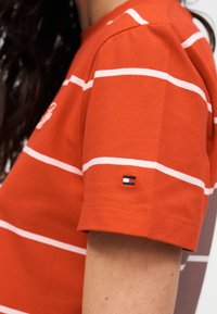 Κόκκινο βαμβακερό μπλουζάκι με λευκές οριζόντιες ρίγες, στρογγυλή λαιμόκοψη και κοντά μανίκια. Διαθέτει μικρό λογότυπο στο μανίκι.