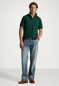 Zelené polo tričko se žlutými akcenty, nošené s světle modrými rovnými džínami. Hnědé boty doplňují outfit. Model stojí před neutrálním pozadím.