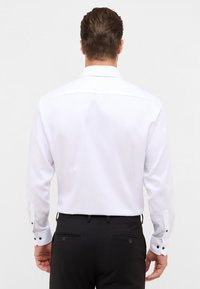 Een man met zijn rug naar de kijker, gekleed in een witte lange mouw overhemd en zwarte formele broek tegen een effen achtergrond.