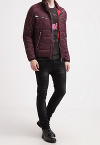 Vínová prošívaná bunda s červenými detaily, kapsami na zip a stojáčkovým límcem, v kombinaci s černými džínami a černými botami.