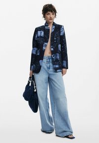 Blazer navy avec des motifs abstraits bleus, associé à un jean large bleu clair. Le mannequin tient un sac navy, mettant en avant un style moderne et décontracté.