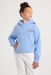 Ragazza con i capelli raccolti in un bun, che indossa un felpa leggera blu chiaro con la scritta "Forever & Always New York", mentre guarda di lato, con una mano in tasca e pantaloni bianchi.