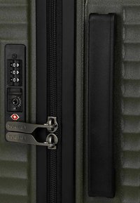 Section de valise texturée verte avec poignée noire, doubles tirettes de fermeture éclair étiquetées "SURI FREY" et un cadenas à combinaison approuvé par la TSA.