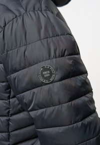 Črno napihnjeno jakno s quiltanim dizajnom. Ima okroglo oznako logotipa U.S. Polo Assn. na zgornjem rokavu. Gladka textura tkanine.