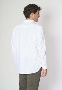 Camicia bianca a maniche lunghe con plissé posteriore e polsini con bottone, abbinata a pantaloni verde oliva. Tessuto morbido con una vestibilità classica.