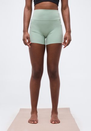 Kvinde står barfodet på en måtte iført lysegrønne atletikshorts med høj talje og en sort crop-top, mod en ensfarvet baggrund.