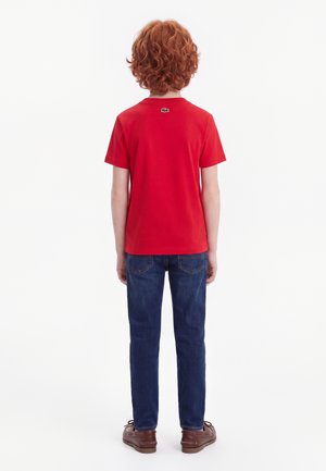 Enfant aux cheveux roux bouclés portant un t-shirt rouge à manches courtes, un jean bleu et des chaussures marron, debout de dos sur un fond blanc.