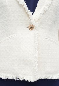 Blazer en tweed blanc avec bords effilochés, un bouton décoratif rond en or, et deux poches avant. Porté sur un vêtement bleu foncé en dessous.