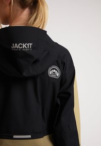 Vattentät jacka med svart huva, beige kropp och en logotyp-etikett. Har en reflekterande detalj vid fållen och en justerbar halsringning.