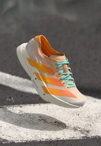Sport shoe in mid-air, featuring an orange and white upper with blue laces and stripes. Background is a blurred road surface.