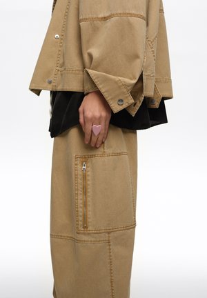 Hand resting by side wearing beige denim jacket and pants with large zippered pocket, pink heart-shaped ring on finger.