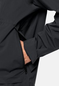 Veste légère noire avec une poche latérale zippée, des poignets ajustables et une texture de tissu lisse ; conçue pour le confort et l'utilité.