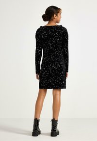 Sort fløjlskjole med lange ærmer, der har hvide stjerne-mønstre. Rynket skulderdetalje og tætsiddende silhuet.