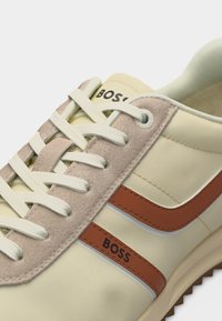 Baskets couleur crème avec zone de laçage en daim beige, bandes latérales marron, lacets blancs et le nom de la marque Boss imprimé sur la languette et le côté.