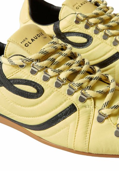Zapatillas de tela amarilla con detalles negros curvos, cordones a rayas, ojales metálicos y la marca "Claudie Paris" en la lengüeta.