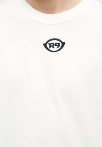 T-shirt blanc à col rond avec petit logo "R9" noir brodé centré sous l'encolure.