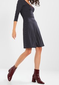 Robe noire à pois blancs avec un décolleté en V et des manches trois quarts. Portée avec des bottines bordeaux brillantes à talons épais.