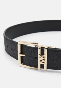 Zwarte leren riem met een gestructureerde afwerking, voorzien van een gouden metalen gesp en logo-details. Verstelbaar met meerdere gaatjes.