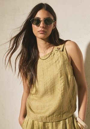 Frau mit langen dunklen Haaren, die ovale Sonnenbrillen trägt, ein ärmelloses hellgelbes Top mit Knopfdetails, eine Kettchenkette und einen passenden Rock.