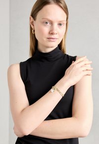 Gold chain bracelet with floral accents and pearl detail, worn on an arm. Black sleeveless top visible, neutral gray background.
