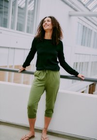 Kvinne med krøllete hår smiler mens hun hviler mot rekkverket i en moderne bygning. Hun har på seg en svart langermet topp, grønne bukser og sandaler.