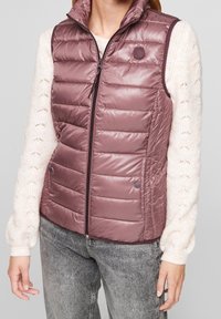 Chaleco ligero sin mangas en color malva, con diseño acolchado, cuello alto y cierre frontal con cremallera. Acentuado con botones de presión en el dobladillo.