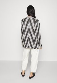 Cardigan tricoté avec un motif chevron noir, blanc et gris. Manches longues, coupe décontractée, avec un col rond, accompagné d'un pantalon blanc à jambes larges.