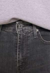 Svarta denimjeans med en slät yta, prydda med en silverfärgad knapp och sömmar. Tuckade under en ljusgrå topp, vilket visar midjebandet.