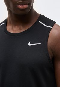 Κοντινό πλάνο ενός ατόμου που φοράει μαύρο αμάνικο αθλητικό μπλουζάκι Nike με λευκό λογότυπο και αντανακλαστικές λεπτομέρειες στους ώμους.