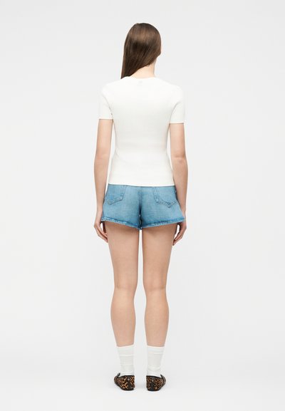 Γυναίκα που στέκεται έχοντας την πλάτη της στραμμένη, φορώντας λευκή κοντομάνικη μπλούζα, ανοιχτό μπλε τζιν σορτς, λευκές κάλτσες και φλατ παπούτσια με λεοπάρ σχέδιο.