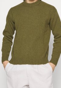 Person som bär en ribbad olivgrön stickad tröja med rund halsringning och ljusbeiga byxor, med händerna i fickorna.