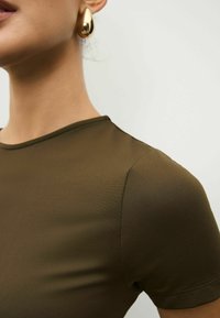 Hnedá priliehavá košeľa s krátkym rukávom a okrúhlym výstrihom, vyrobená z hladkého materiálu, s jemnými švami na pleciach. Zlaté náušnice v tvare slzy.