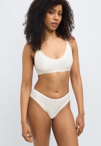 Λευκή σουτιέν με διακοσμητική δαντέλα και ασορτί σλιπ, που διαθέτει υφή με λεπτομερή σχέδια και μικρές φιόγκους ως διακοσμητικά στοιχεία.