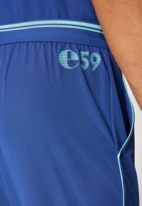 Blaue Sportshorts aus glattem Stoff, mit einer Netzstruktur, einem hellblauen Streifen am Bund und einem aufgedruckten "c59" Logo.