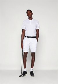 Hvit poloskjorte med krage, kombinert med hvite shorts. Svart belte og svarte joggesko fullfører antrekket. Holder en golfkølle.