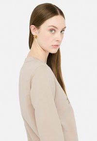Maglietta beige a maniche lunghe con scollatura tonda. Tessuto leggero e liscio con dettagli di cucitura discreti. Il modello posa di lato, mostrando accessori minimi.