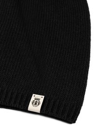 Black knit beanie with ribbed texture, featuring a light-colored tag with a logo at the base.