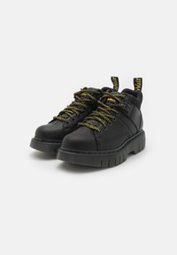 Sapatos de couro pretos com superfície texturizada, sola grossa preta e atacadores com detalhes amarelos. Apresenta uma aba proeminente no calcanhar para facilitar o calçar.