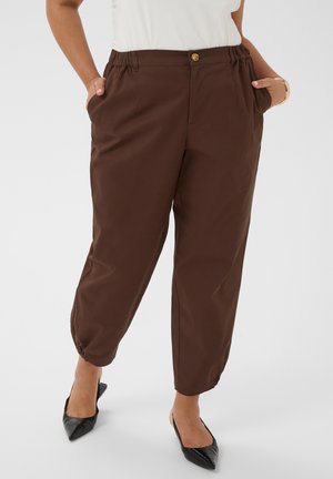 Žena nosi smeđe hlače do gležnja s elastičnim manšetama i crne šiljate balerinke, ruke u džepovima, stoji na bijeloj pozadini.