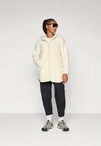 Light beige fleece jacket with a front zipper, hood, and side pockets, paired with dark pants and grey athletic shoes. Shiny sunglasses worn.
