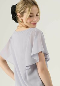 Donna bionda con orecchini e collana d'oro, che indossa una blusa leggera grigia con maniche fluide, sorridente e che guarda in basso, con un accessorio per capelli nero.