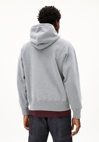 Mann trägt eine hellgraue Kapuzenjacke über einem kastanienbraunen Hemd und dunkler Hose, steht mit dem Rücken zu uns vor einem schlichten weißen Hintergrund.