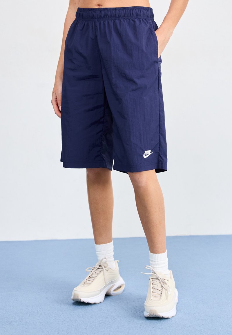 Personne debout en short de sport Nike bleu marine, les mains dans les poches, avec des chaussettes blanches et des baskets beiges sur un sol bleu clair.