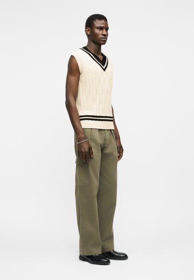 Νεαρός άνδρας που στέκεται, φοράει αμάνικη μπεζ πλεκτή μπλούζα με μαύρες ρίγες, χακί παντελόνια με τσέπες cargo, μαύρα παπούτσια και ασημένιο βραχιολάκι.