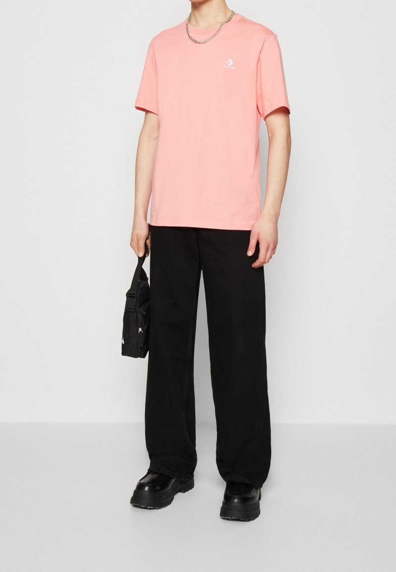 Personne portant une chemise rose à manches courtes, un pantalon large noir, des chaussures noires épaisses, un collier chaîne en argent, tenant un petit sac noir.