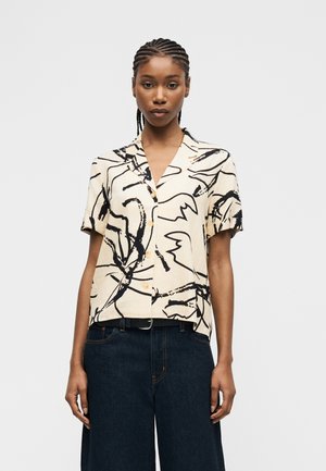 Femeie cu păr împletit, purtând o bluză crem cu mâneci scurte și imprimeu abstract negru și pantaloni înalți, largi, de culoare închisă.