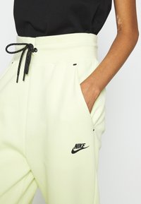 Light yellow sweatpants with a black drawstring, side pockets, and a black Nike logo on the thigh. Smooth fabric texture.