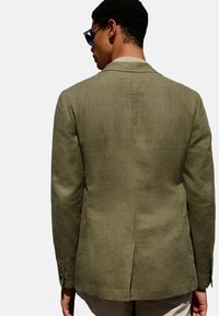 Olivenfarbener Leinenblazer mit taillierter Passform, einem Schlitz hinten und subtiler Texturfläche. Goldene Knöpfe an den Ärmeln.