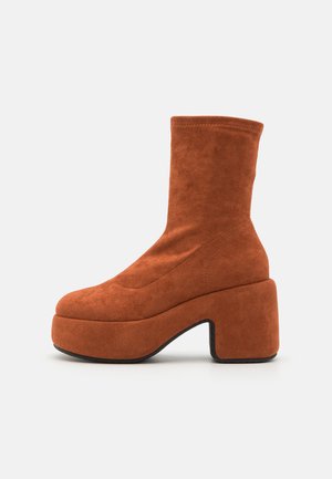 Καφέ σουέτ μποτάκια μέχρι τον αστράγαλο με τακούνι τύπου block και πλατφόρμα. Το επάνω μέρος διαθέτει λείο υλικό και λεπτομέρειες ραφής, φτάνοντας μέχρι το μέσο της γάμπας.