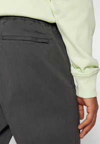 Donkergrijze broek met een gladde textuur, voorzien van een achterzak en een elastische tailleband, gecombineerd met een lichtgroene trui.